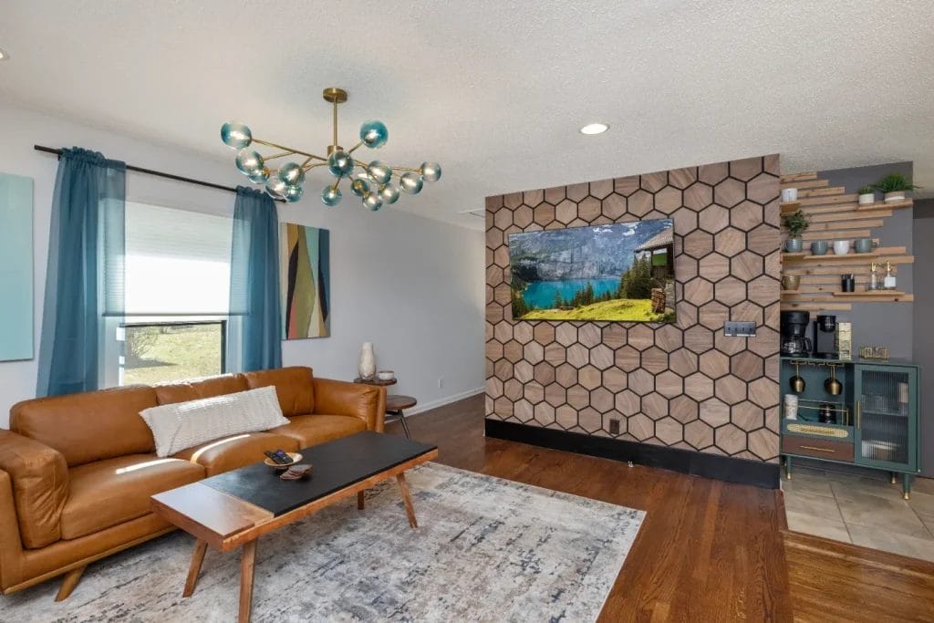 A modern living room with a brown leather sofa, a wooden coffee table, a geometric rug, and a wall-mounted TV displaying a nature scene. The accent wall features hexagonal wood tiles and open shelves with decor.