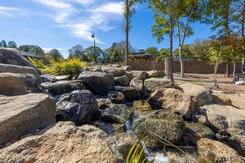 A small stream with clear water flows over large rocks surrounded by trees and greenery, under a blue sky with scattered clouds in a landscaped outdoor area.