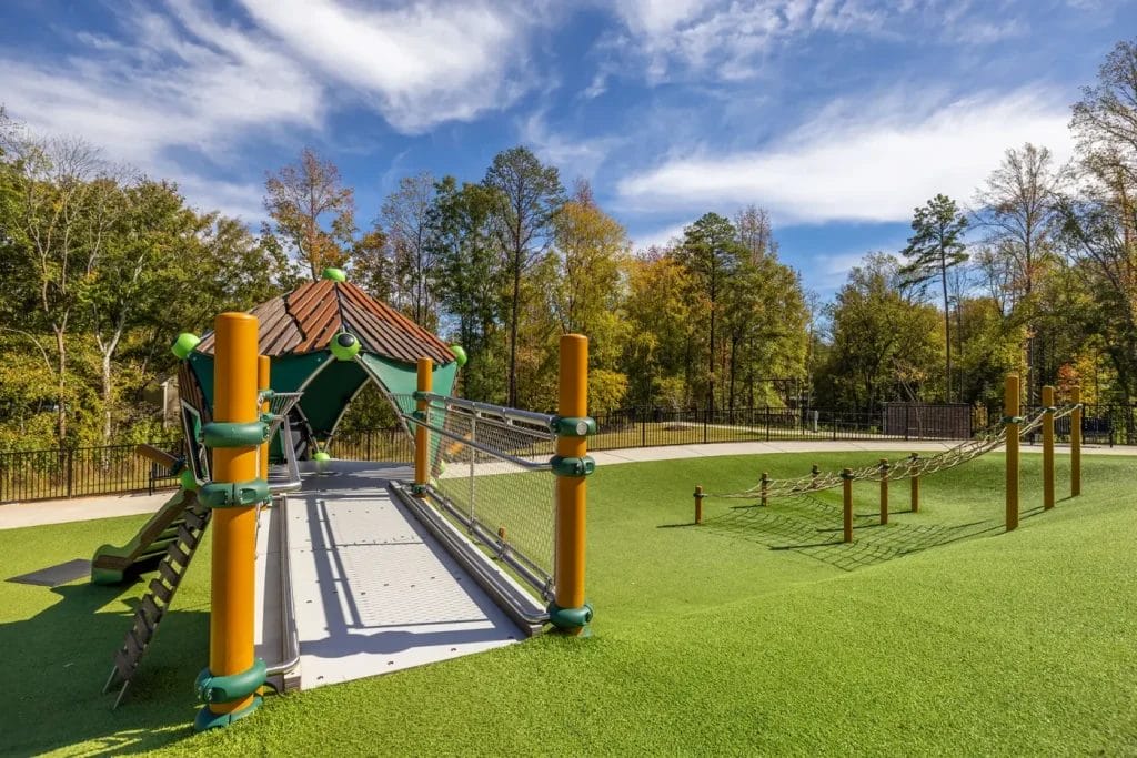 A modern playground with a green and yellow climbing structure, a bridge, rope obstacles, and soft artificial grass, surrounded by trees under a partly cloudy blue sky.