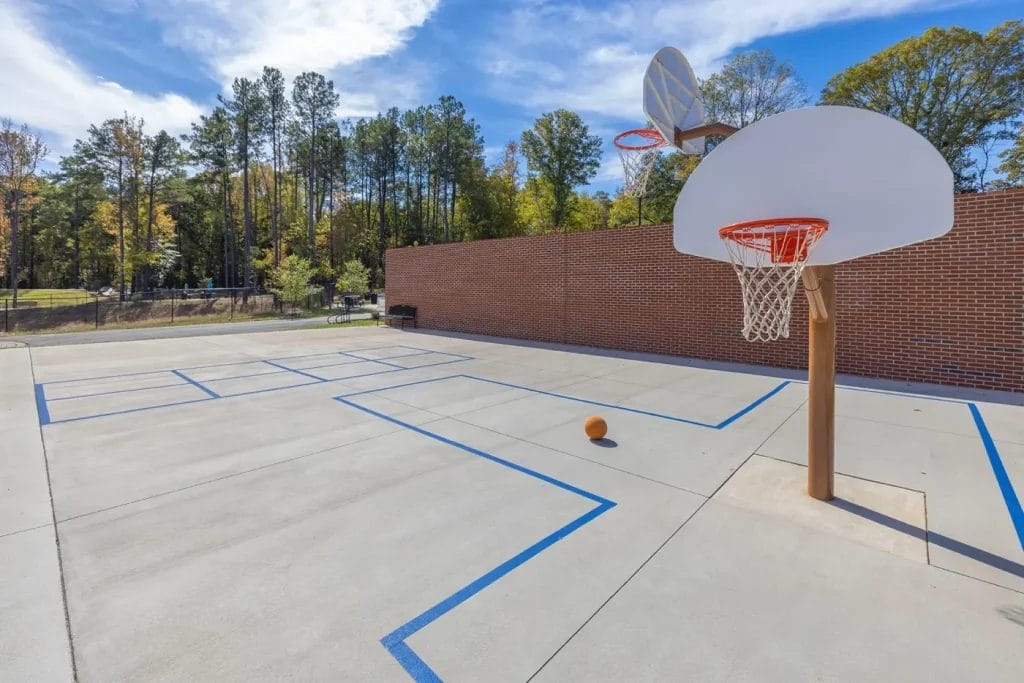 An outdoor basketball court with blue lines on concrete, a basketball hoop and backboard in the foreground, a basketball on the court, a brick wall, trees, and a blue sky with clouds in the background.