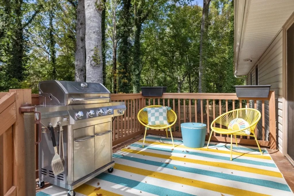 A backyard deck with a stainless steel grill, two yellow chairs, a blue side table, and a striped outdoor rug. The area is enclosed by a wooden railing and surrounded by trees.