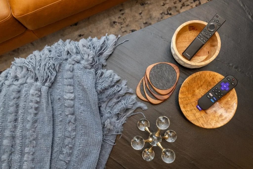 A dark coffee table with two wooden bowls holding remote controls, a set of coasters, a decorative piece, and a textured gray throw blanket draped nearby. Part of a tan sofa and a patterned rug are also visible.