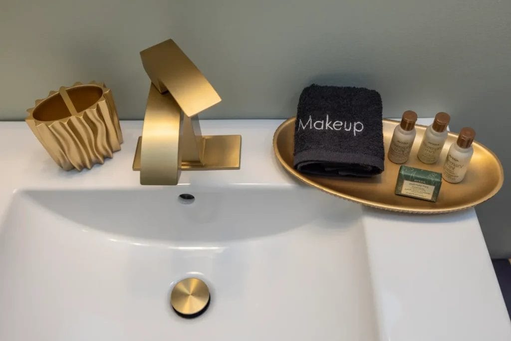 A modern bathroom sink with a gold faucet, a decorative gold cup, and a gold tray holding toiletries, soap, and a black towel labeled "Makeup." The countertop and sink are white.