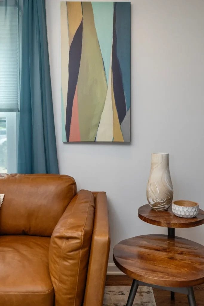 A tan leather sofa sits beside two round wooden side tables with ceramic vases and a small bowl. An abstract, colorful painting hangs on the wall above, and a blue curtain frames a window to the left.
