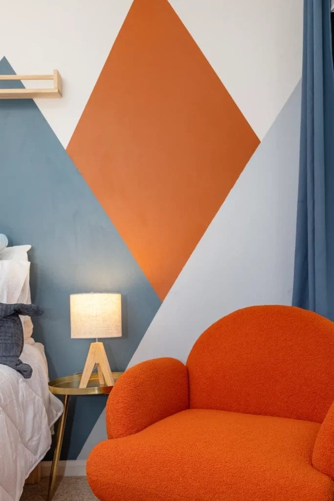 A cozy orange armchair sits beside a small round table with a lamp, in front of a wall with geometric shapes in orange and blue behind a white bed.