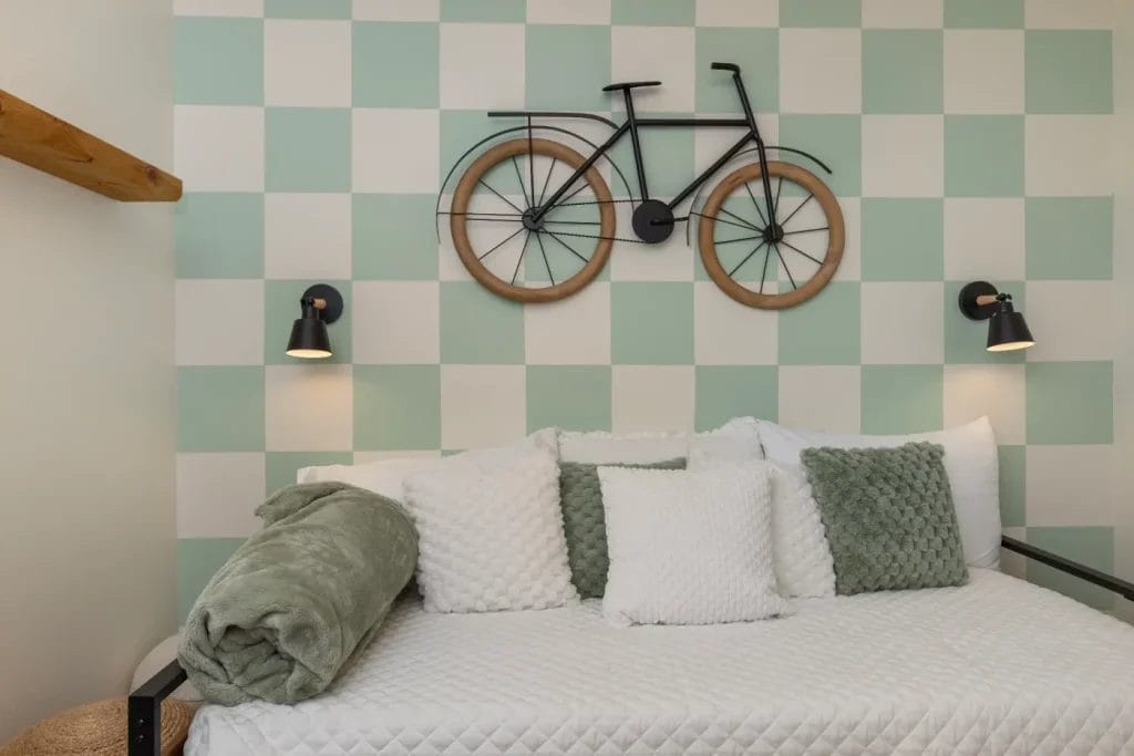 A bed with white and green textured pillows and a rolled green blanket sits against a green-and-white checkered wall. Above the bed hangs a decorative bicycle art piece and two black wall-mounted reading lamps.