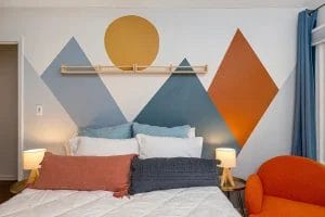 A cozy bedroom with a bed featuring blue, white, and rust-colored pillows, flanked by two lamps. The wall behind the bed is decorated with geometric mountain shapes and a sun in orange and blue tones. An orange chair sits nearby.