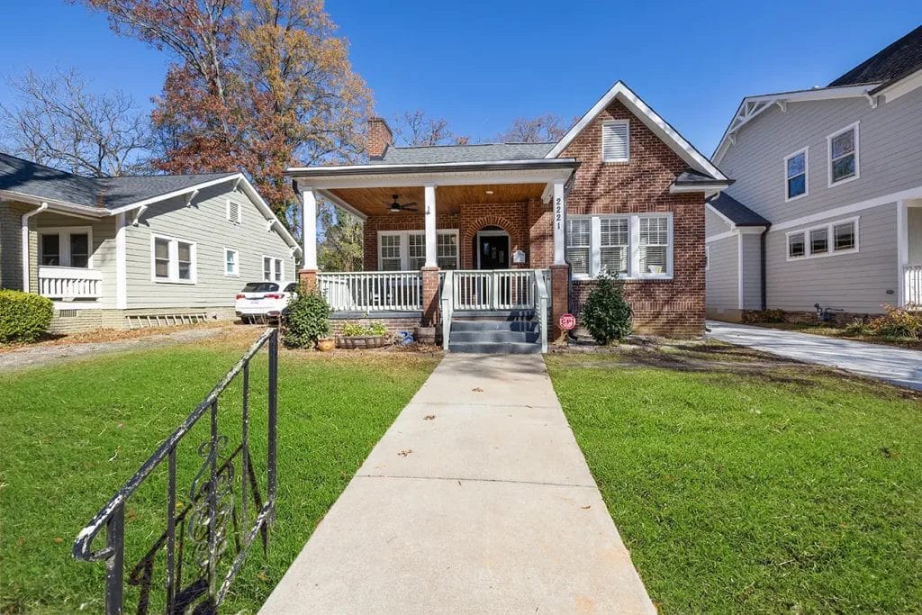 A brick house with a covered front porch, white railings, and steps leading up from a concrete walkway. The house is surrounded by a green lawn and neighboring homes under a clear blue sky.