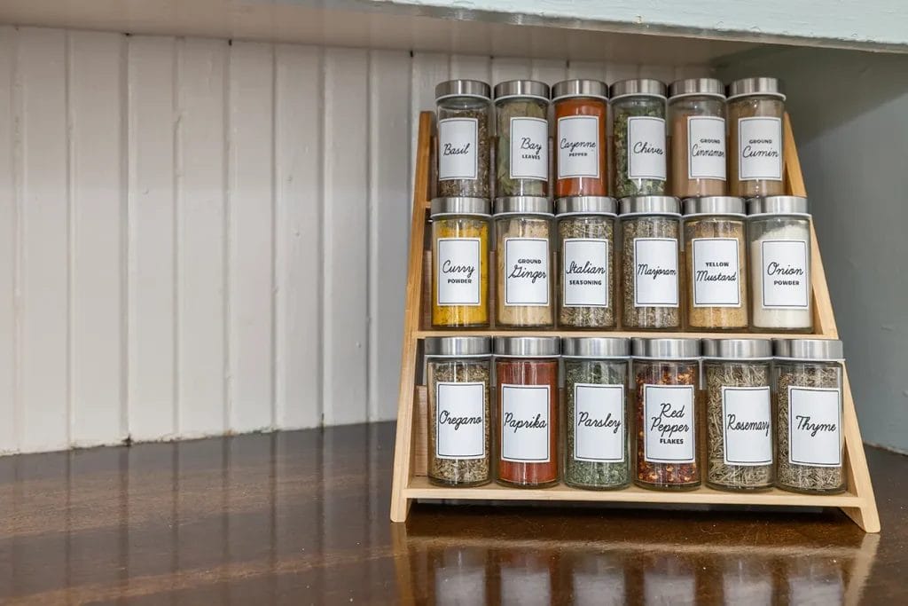 A wooden spice rack holds three rows of glass jars with metal lids, each labeled with a different spice such as basil, cayenne, oregano, thyme, parsley, rosemary, and cinnamon, arranged neatly on a kitchen counter.