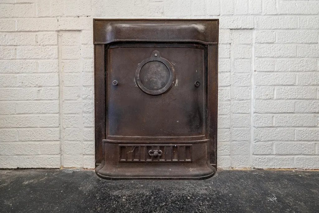 A vintage metal furnace or stove door is mounted in a white-painted brick wall, with a black stone or concrete floor beneath it. The door has a circular vent and decorative details.
