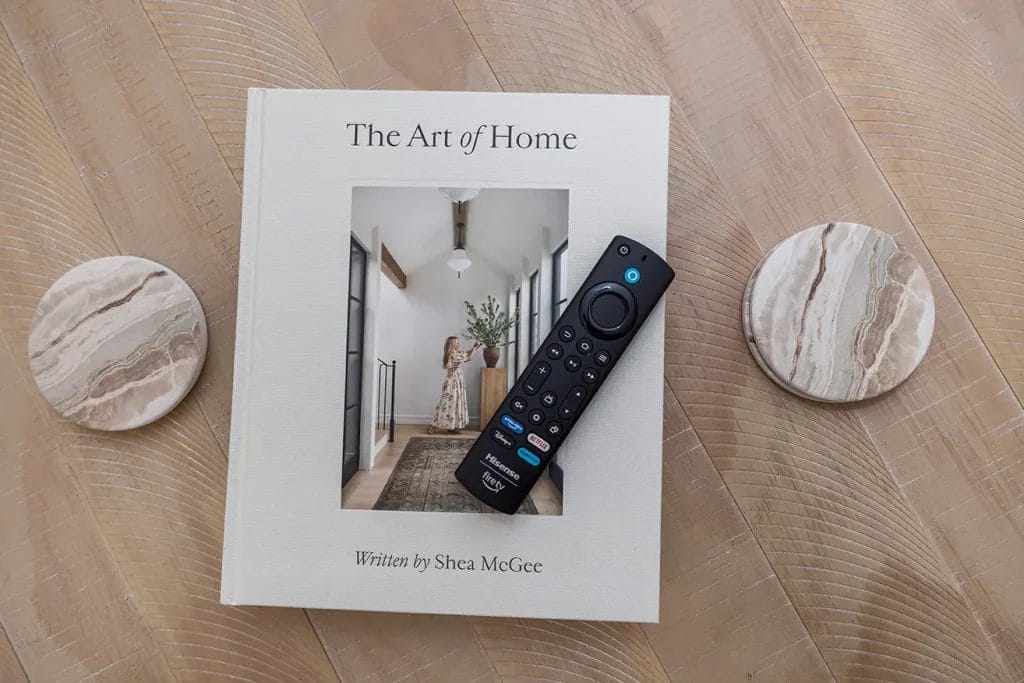 A book titled "The Art of Home" by Shea McGee lies on a wooden surface, with a TV remote placed on top. Two round stone coasters are positioned on either side of the book.
