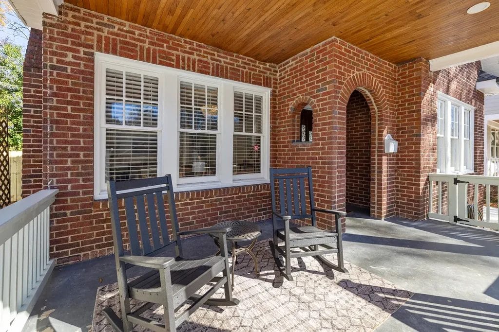 Two black rocking chairs and a small table sit on a covered porch with a brick wall, white-trimmed windows, and a wooden ceiling. Sunlight streams in, creating a cozy, inviting outdoor space.