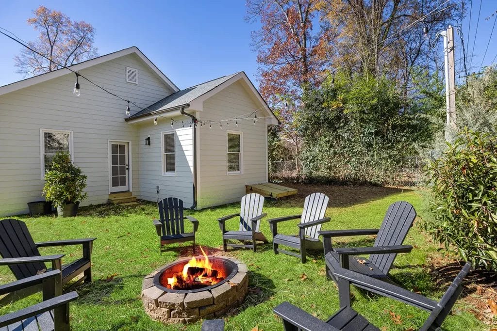 A backyard with a white house, string lights, and Adirondack chairs arranged around a stone fire pit on green grass, with tall trees and bushes in the background.