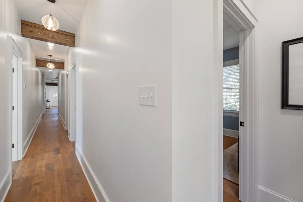 A bright hallway with white walls, wood flooring, modern light fixtures, and several doorways, including one leading to a room with a window and blinds.