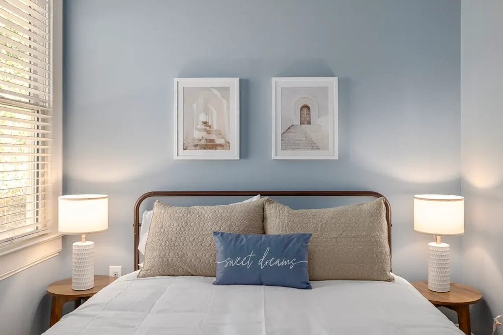 A neatly made bed with beige pillows and a blue cushion reading "sweet dreams," flanked by two wooden nightstands with white lamps, against a light blue wall with two framed architectural photos above the headboard.