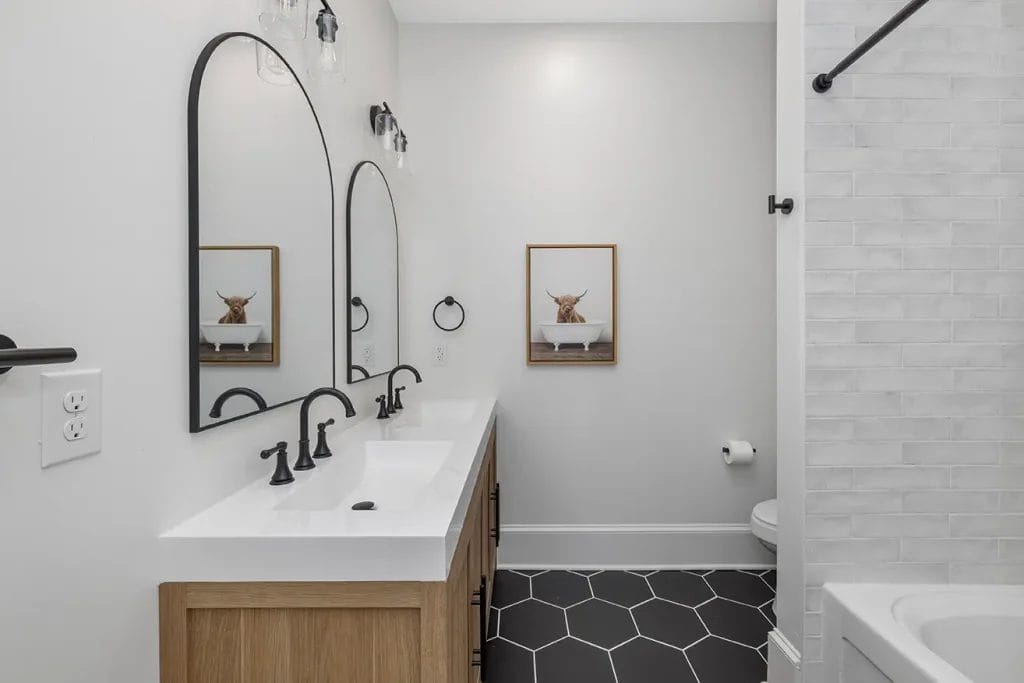 Modern bathroom with double sink vanity, black fixtures, two arched mirrors, hexagonal black floor tiles, white walls, a toilet, a bathtub with white tile, and framed art of a cow above the toilet.