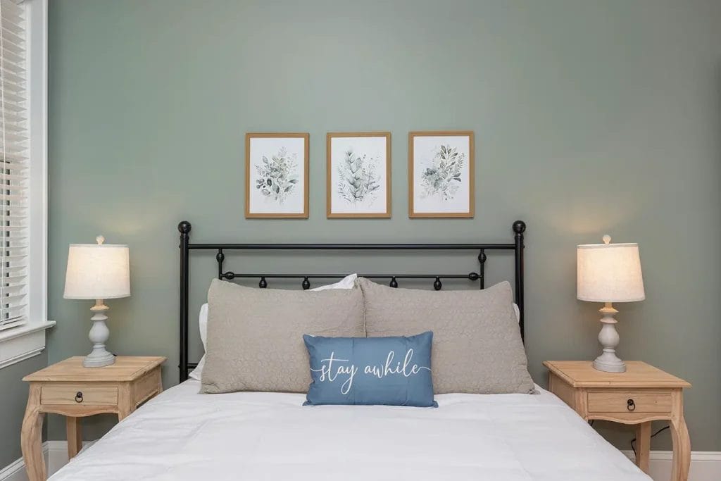 A neatly made bed with gray pillows and a blue cushion that says "stay awhile" sits between two wooden nightstands with lamps. Three framed botanical prints hang above the bed against a green wall.