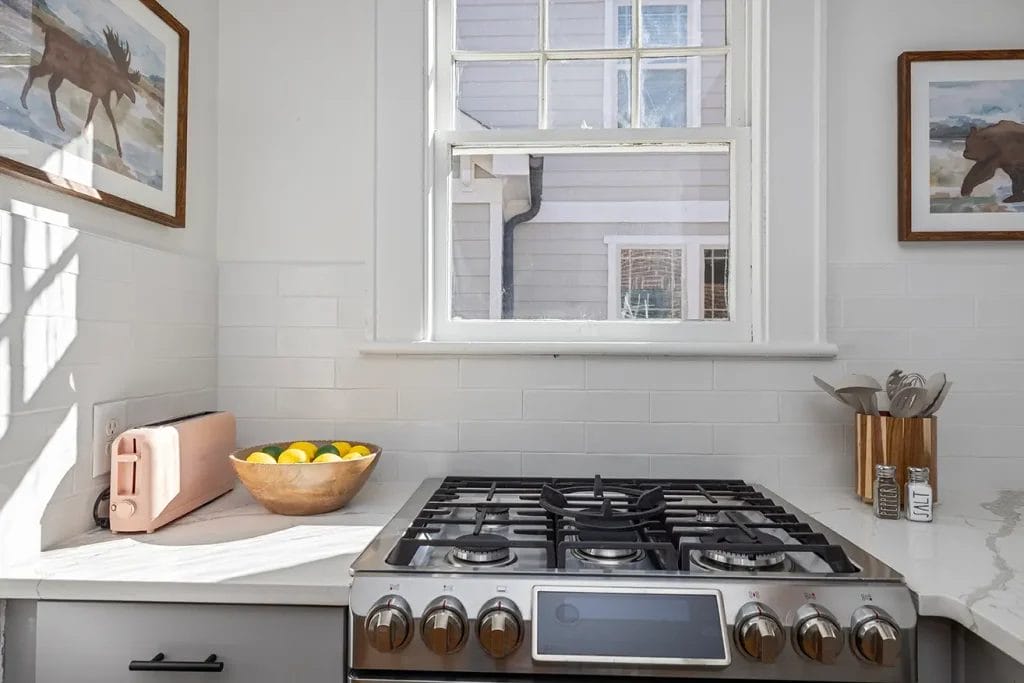 A kitchen with a gas stove, a bowl of lemons, a pink toaster, and utensils on a white countertop. There are animal paintings on the walls and a window above the stove letting in natural light.