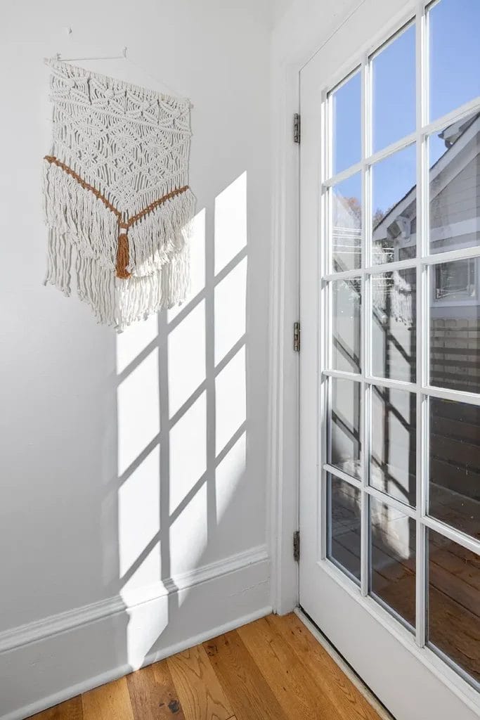A glass-paneled door lets sunlight into a corner with light-colored walls, casting shadows on the wall below a decorative white macramé wall hanging with fringe and a wooden accent. The floor is light wood.