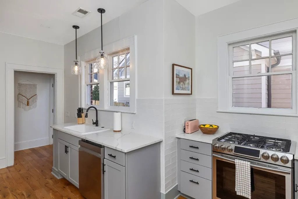 A bright, modern kitchen with light gray cabinets, white countertops, a farmhouse sink, stainless steel dishwasher, gas stove, pink toaster, bowl of lemons, and two pendant lights above the sink.