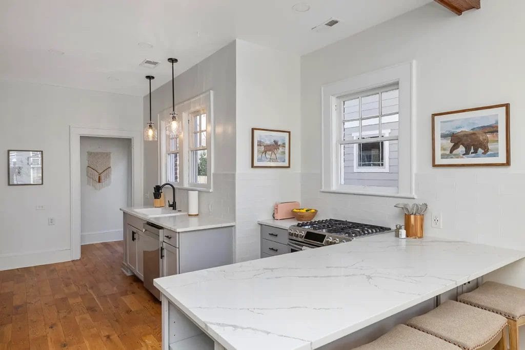 Modern kitchen with white cabinets, marble countertops, stainless steel appliances, pendant lights, and wooden flooring. Two framed animal pictures and a window are on the white walls. Three stools are at the counter.
