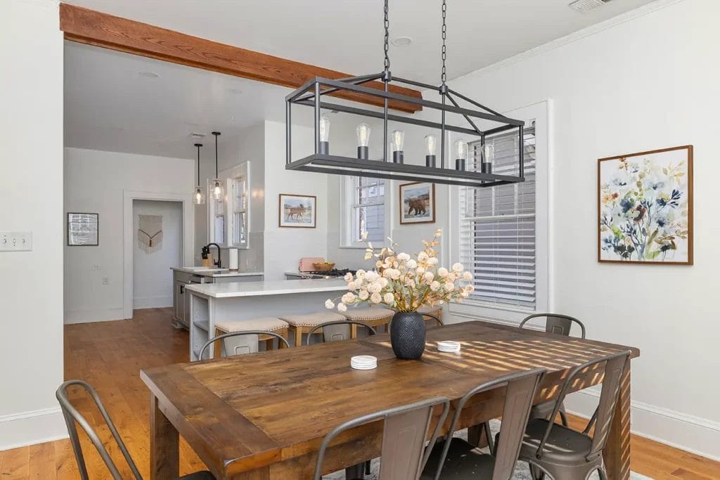 Modern dining room with a wooden table, six metal chairs, a black geometric chandelier, and a vase of white flowers. The space opens to a kitchen with white cabinets and pendant lights. Light streams in through large windows.