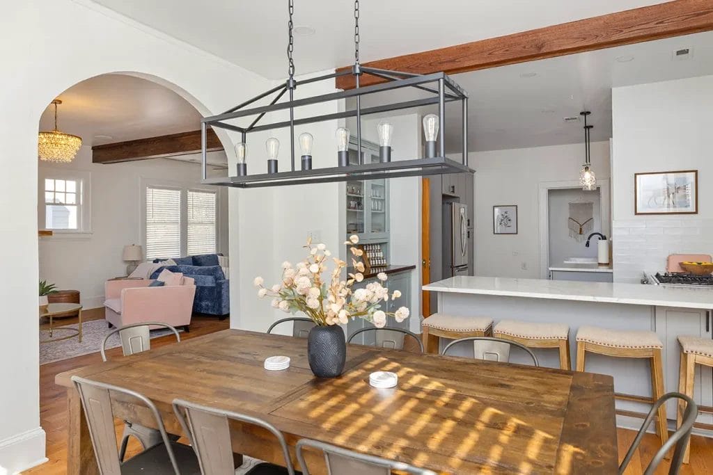 A modern dining room with a wooden table, metal chairs, a black rectangular chandelier, and a vase of flowers. The kitchen with bar stools and a living room with sofas are visible in the background.