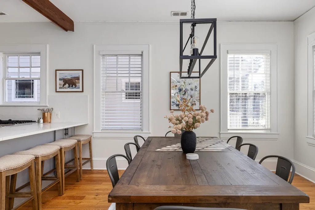 A bright dining area with a wooden table, black chairs, a black geometric light fixture, and a vase of dried flowers. Three windows let in natural light, and barstools line a white counter to the left. Art hangs on the white walls.
