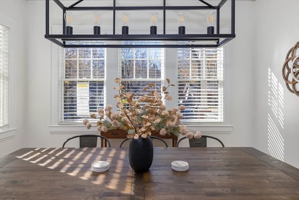 A wooden dining table with a black vase holding dried flowers sits beneath a modern rectangular chandelier. Light streams through large windows with white blinds, casting shadows on the table.