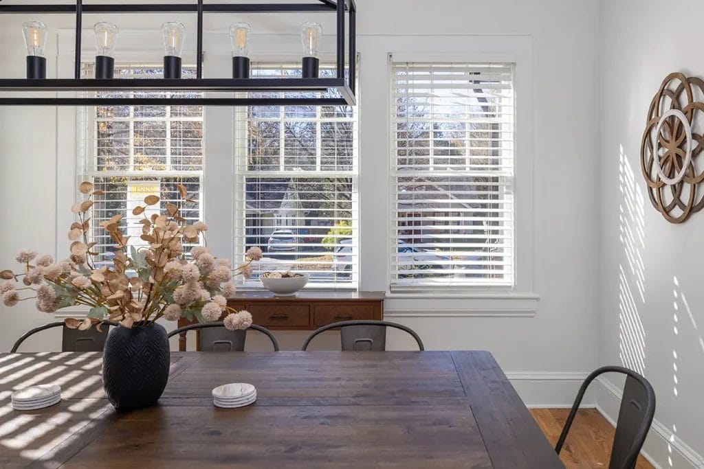 A bright dining room with large windows, white blinds, a wooden table, metal chairs, a black vase with dried flowers, wall art, and a modern light fixture. Sunlight streams in, casting shadows on the floor and table.