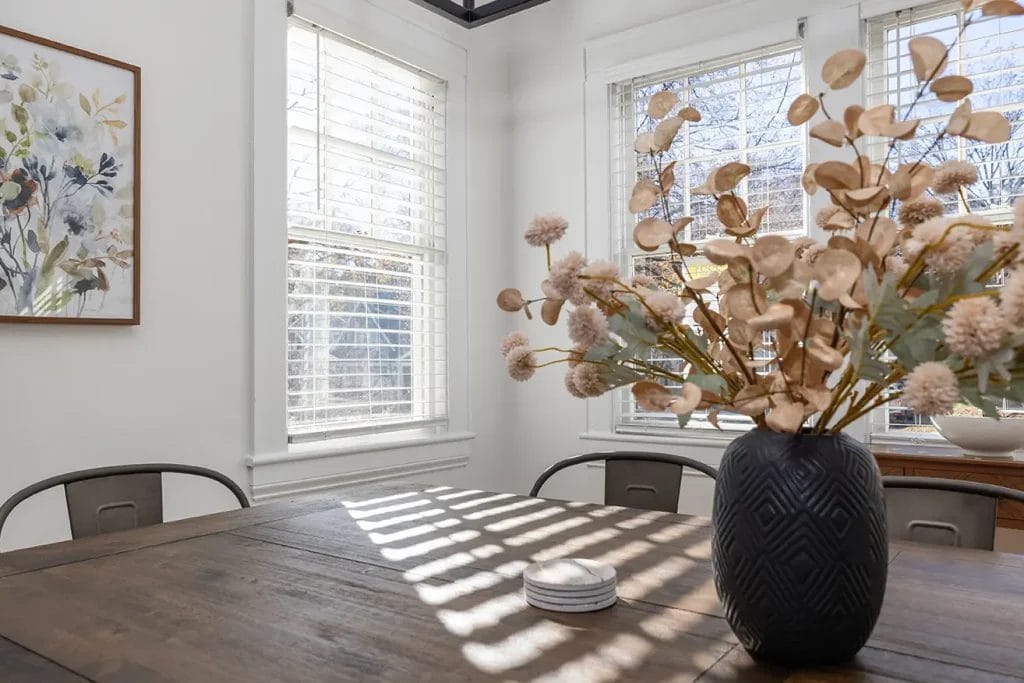 A sunlit dining room with a wooden table, black vase holding dried flowers, metal chairs, and large windows casting striped shadows. A floral painting hangs on the white wall.