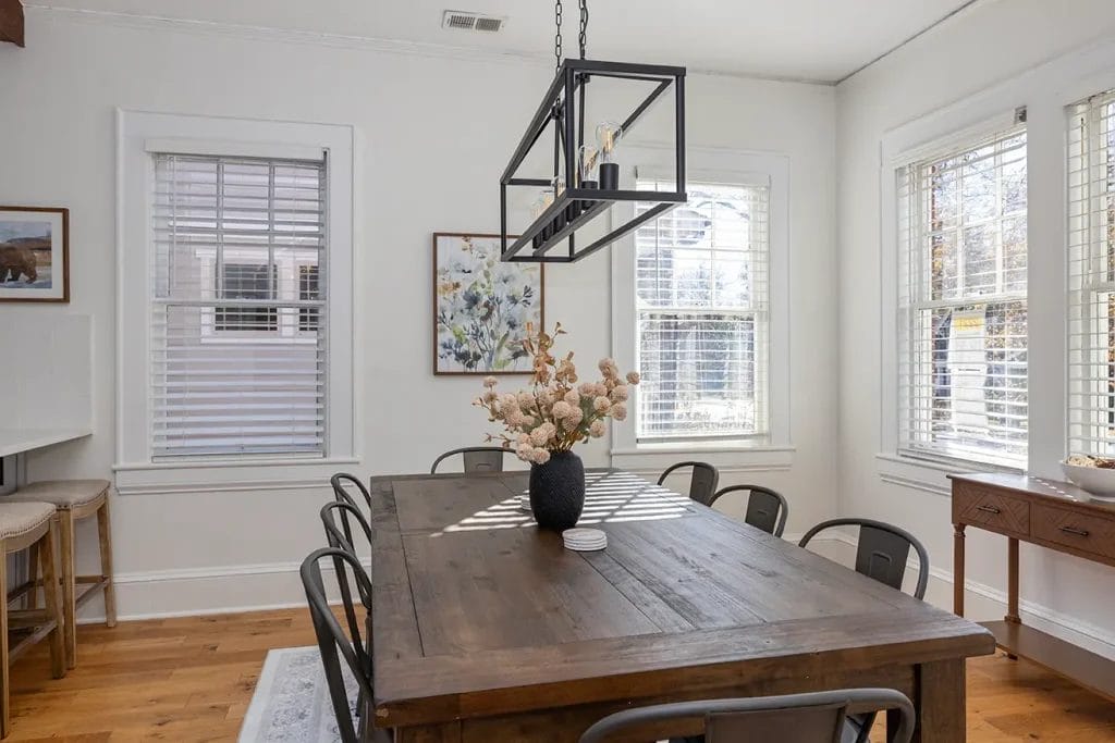 Bright dining room with a wooden table, eight metal chairs, a black overhead light fixture, and a vase with flowers. Large windows with white blinds let in natural light, and framed art hangs on the white walls.
