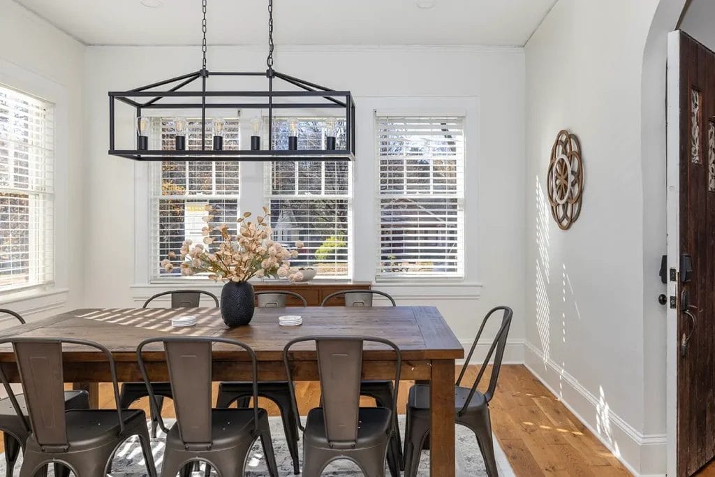 A bright dining room with a large wooden table, eight metal chairs, a modern rectangular chandelier, a black vase with dried flowers, wooden floors, and white walls with windows letting in natural light.