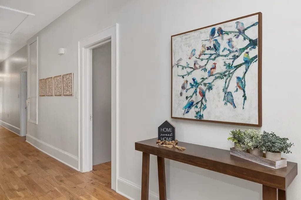 A hallway with wooden floors, white walls, and a wooden console table decorated with potted plants and a small "Home Sweet Home" sign. A large bird painting hangs above the table. Three small artworks are on the far wall.