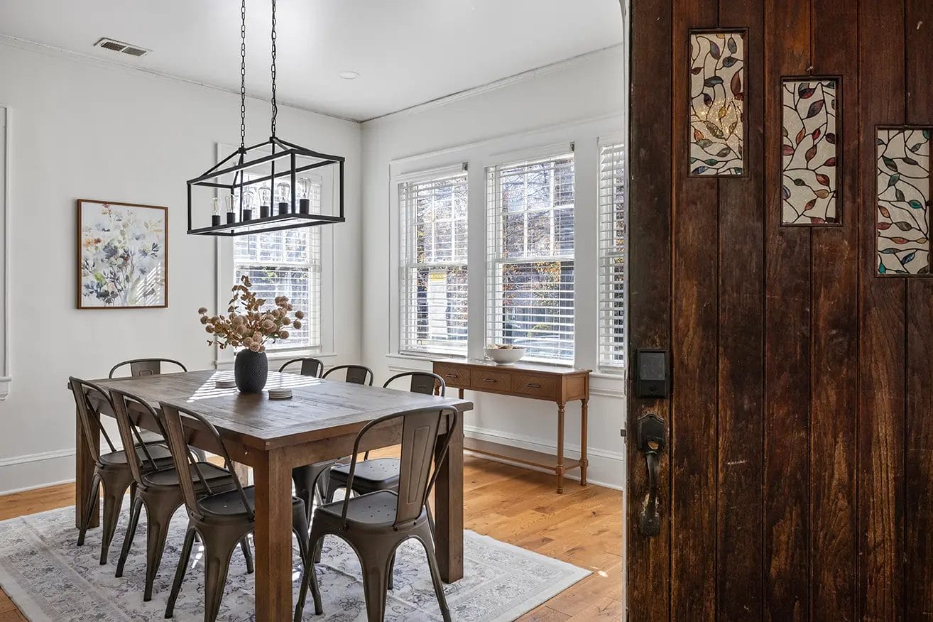 A dining room with a wooden table set for six, metal chairs, a modern black chandelier, a rug, a side table under two large windows, and artwork on the wall. Part of a rustic wooden door is visible in the foreground.