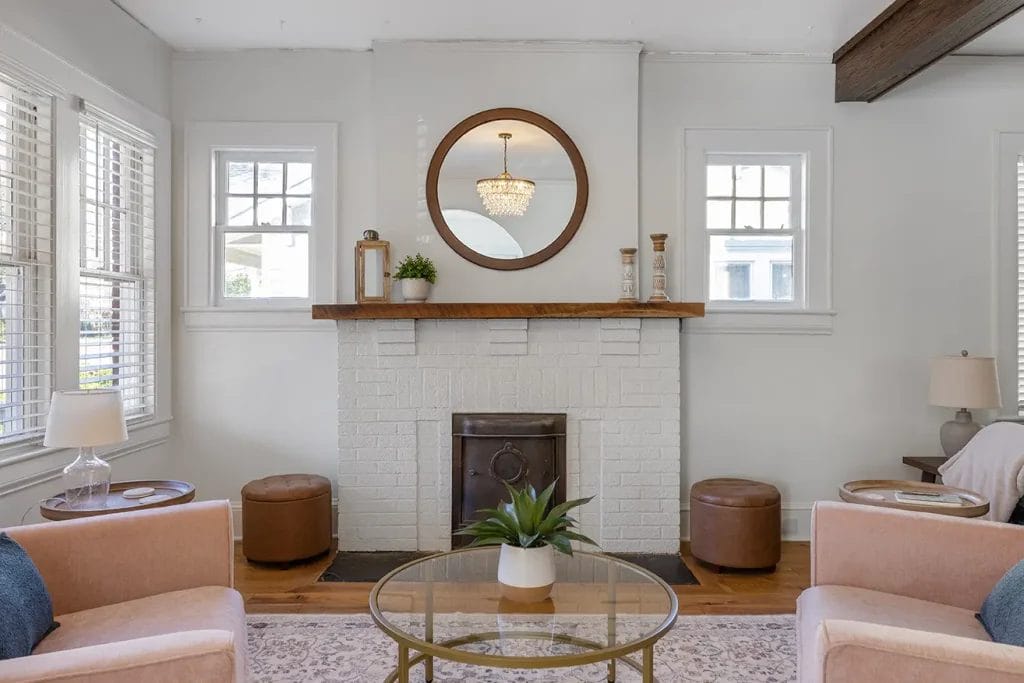 A cozy living room with a white brick fireplace, round mirror, and wooden mantel. Two brown ottomans sit on either side, with beige armchairs and a glass coffee table with a potted plant in the foreground.