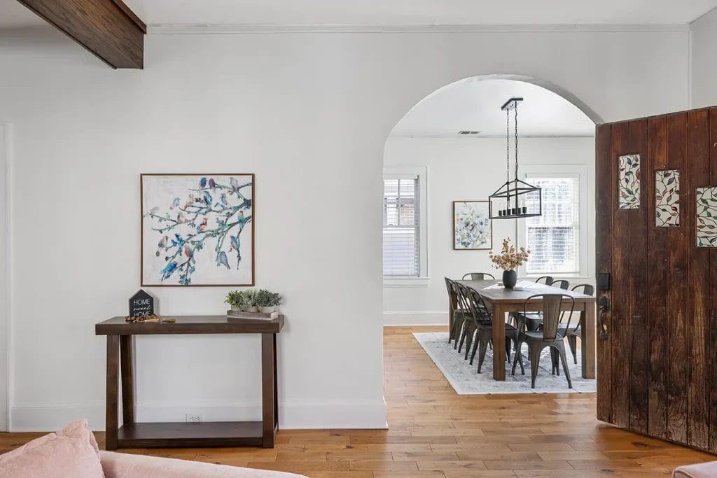 A bright dining room with a long wooden table and metal chairs is visible through an arched doorway. In the foreground, a console table with decor and artwork hangs on a white wall. The floor is light wood.