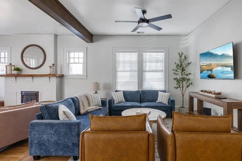 Modern living room with two blue sofas, two brown leather chairs, a round coffee table, wall-mounted TV, fireplace with a circular mirror, potted plant, and wood beams on the white ceiling. Natural light fills the space.