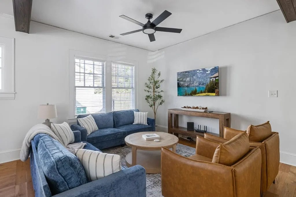 A modern living room with two blue sofas, two brown leather chairs, a round wooden coffee table, a TV mounted on the wall, a console table, and a large window letting in natural light.