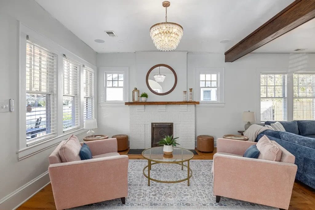 Bright living room with two pink armchairs, a blue sectional sofa, a round glass coffee table, and a white brick fireplace with a wooden mantel and round mirror. Large windows let in natural light.