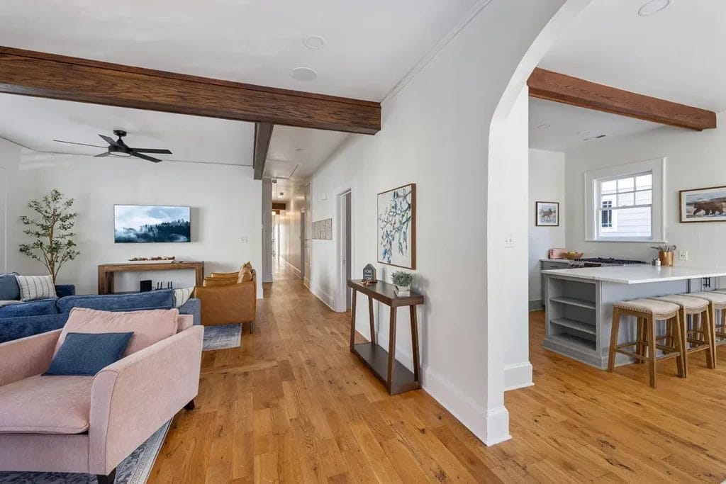 Bright open living space with wood floors, a cozy seating area, a wall-mounted TV, decorative beams, and an archway leading to a modern kitchen with a breakfast bar and stools.