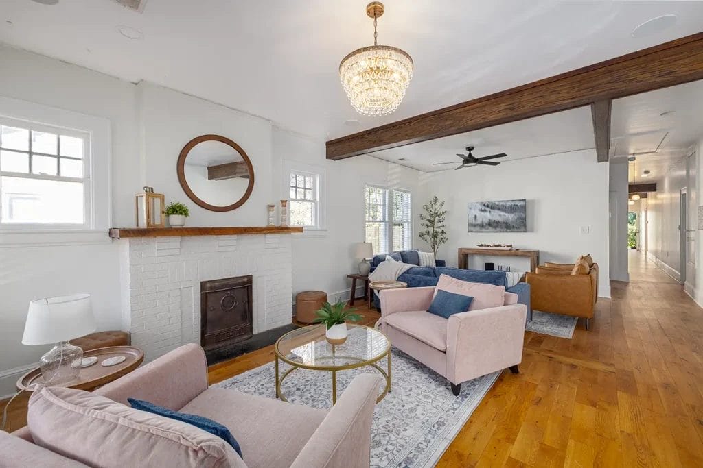 Bright living room with hardwood floors, neutral-toned furniture, a white brick fireplace with a round mirror above, a chandelier, and large windows letting in natural light. Exposed wooden beams add a rustic touch.