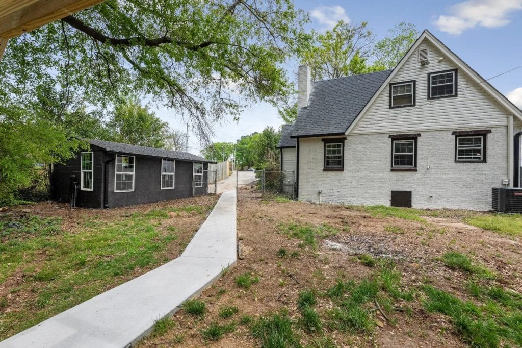 A concrete path runs through a grassy yard, leading to a black shed and a white brick house with gray roof shingles, surrounded by trees and a wooden fence in the background.