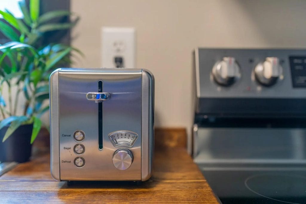 A silver toaster with buttons and a dial sits on a wooden kitchen counter next to a stove. A green plant and an electrical outlet are visible in the background.