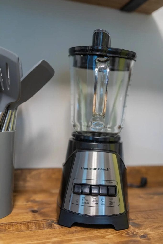A Hamilton Beach blender with a clear glass pitcher sits on a wooden countertop next to a utensil holder with kitchen tools. The blender has several black and gray control buttons on its front panel.