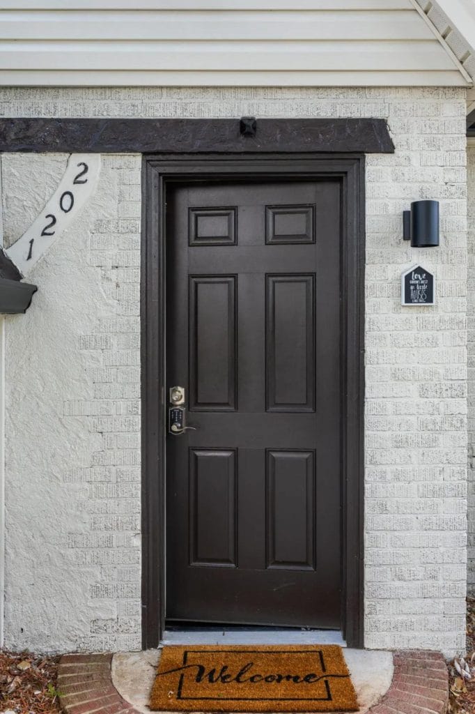 A black front door with a "Welcome" doormat, house number "1202" on the left, and a small sign reading "Love Lives Here" mounted on the white brick wall beside the door.
