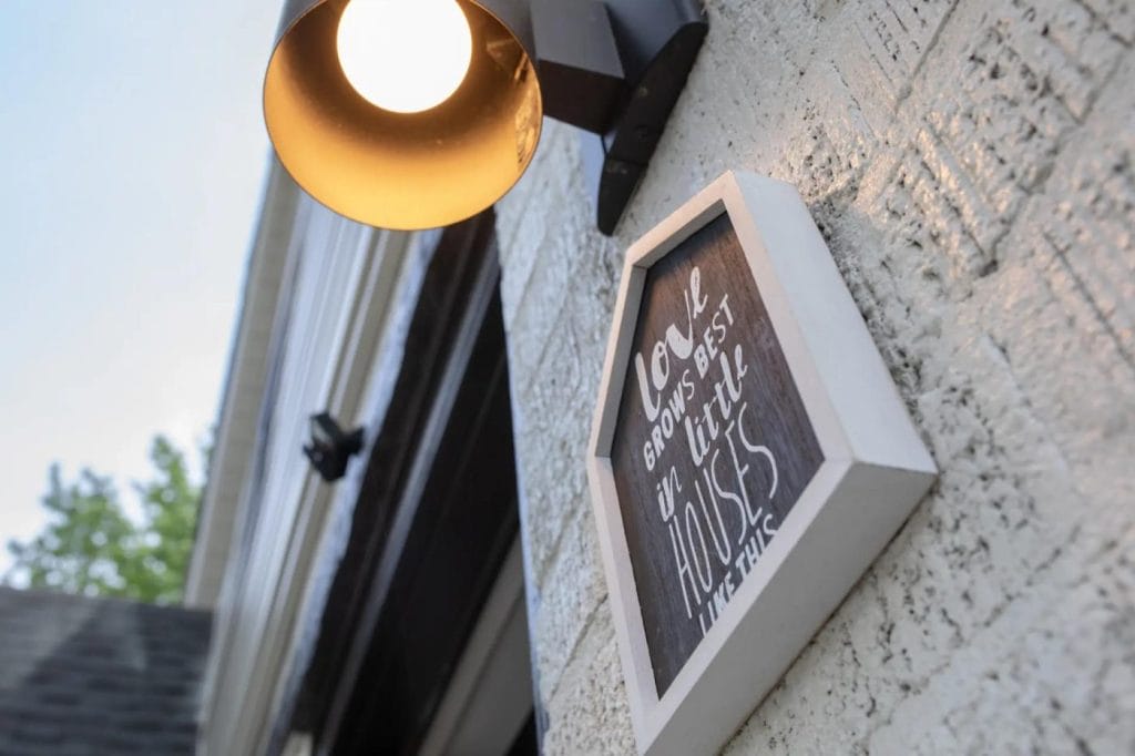 A wall light illuminates a small framed sign on a textured white brick wall. The sign reads, “Love grows best in little houses like this.” A hint of trees and blue sky are visible in the background.