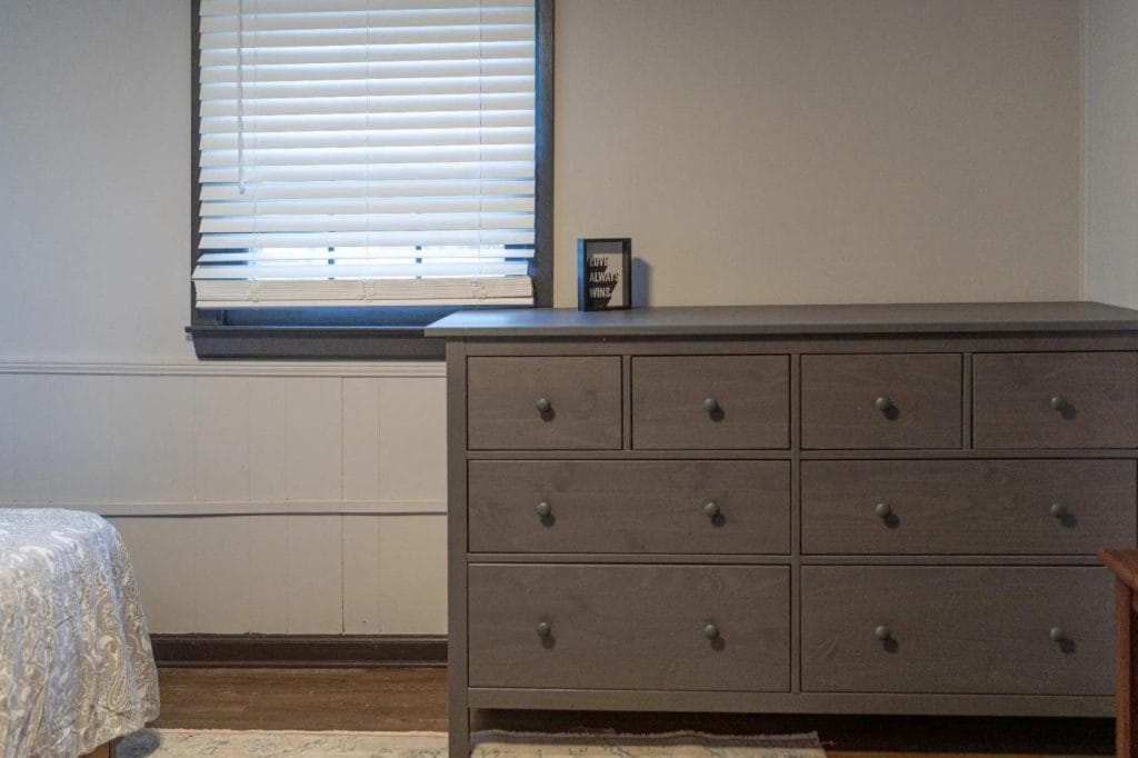 A gray dresser with nine drawers stands against a light-colored wall beneath a window with closed blinds. A small decorative sign sits on top of the dresser. The corner of a bed is visible on the left.