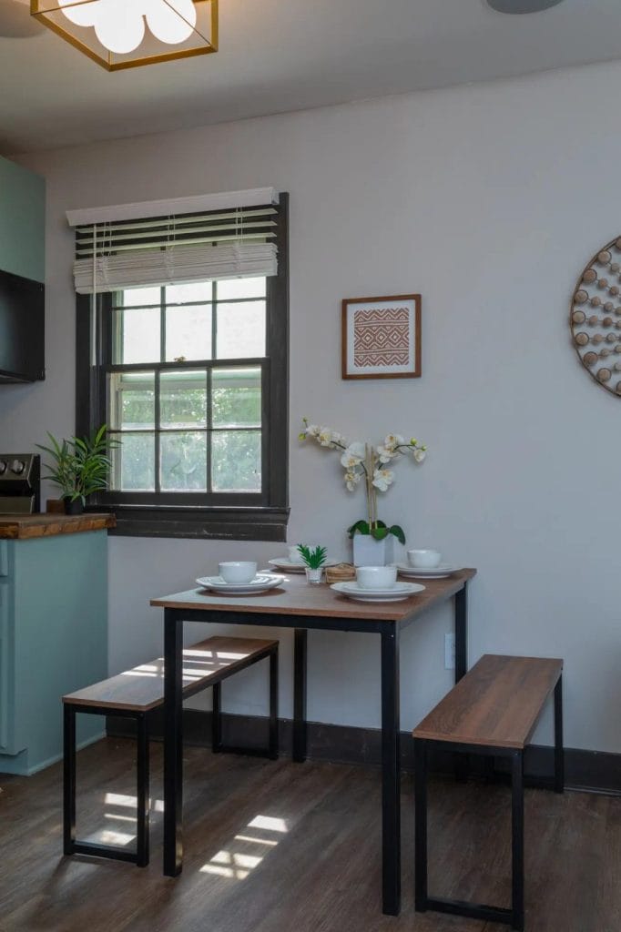 A small dining area with a wooden table, two benches, place settings, a potted orchid, and wall decor. Sunlight enters through a window with blinds, and a green plant sits on the counter nearby.