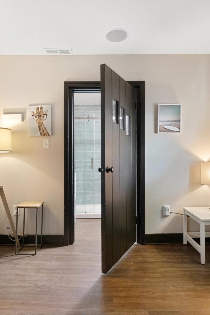 A partially open dark brown door reveals a bathroom with a glass shower. The room has wood flooring, beige walls, framed art, two lamps, and simple furniture including a side table and desk.